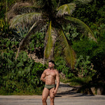 Man standing shirtless in a tropical setting with palm trees and greenery, wearing Caio's Astro Moss Classic Swim Brief. 