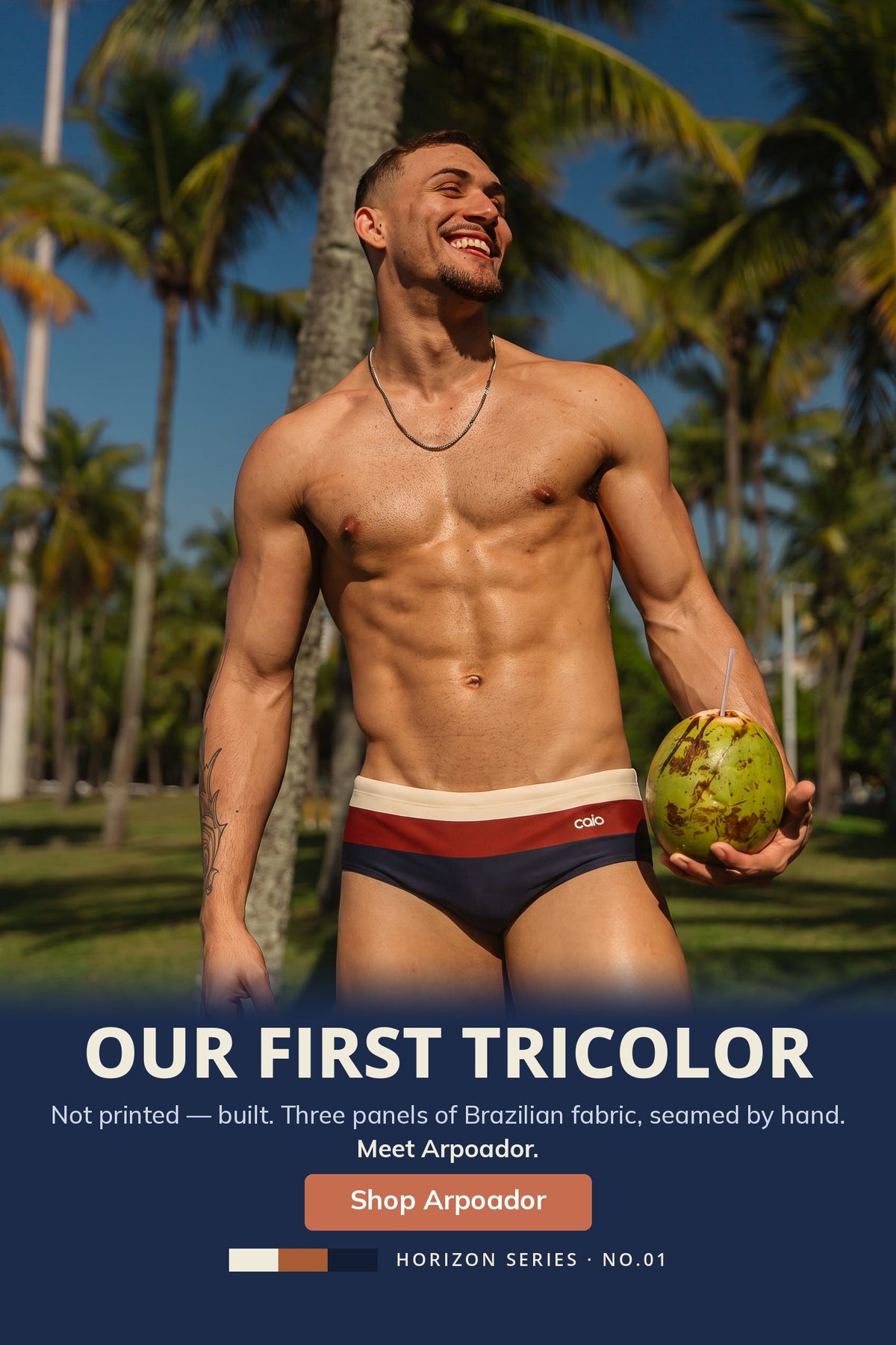 A shirtless man outdoors among palm trees wears tricolor swim briefs with a cream, navy, and red horizontal stripe pattern and holds a coconut. Large text at the bottom reads 'OUR FIRST TRICOLOR' and 'SHOP ARPOADOR'.
