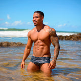 Model standing in shallow water at the beach wearing a Caio Classic Stellar Ash Swim Brief, with a clear blue sky in the background.