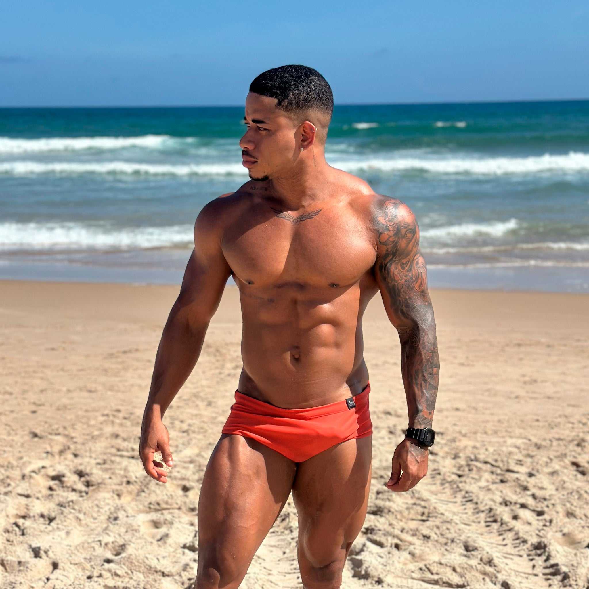 Model wearing Abreu Classic Nina Sunga Swim Brief standing on a beach with ocean waves in the background