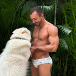 man in caio slim nova frost swim brief with a white dog against green leafy backdrop