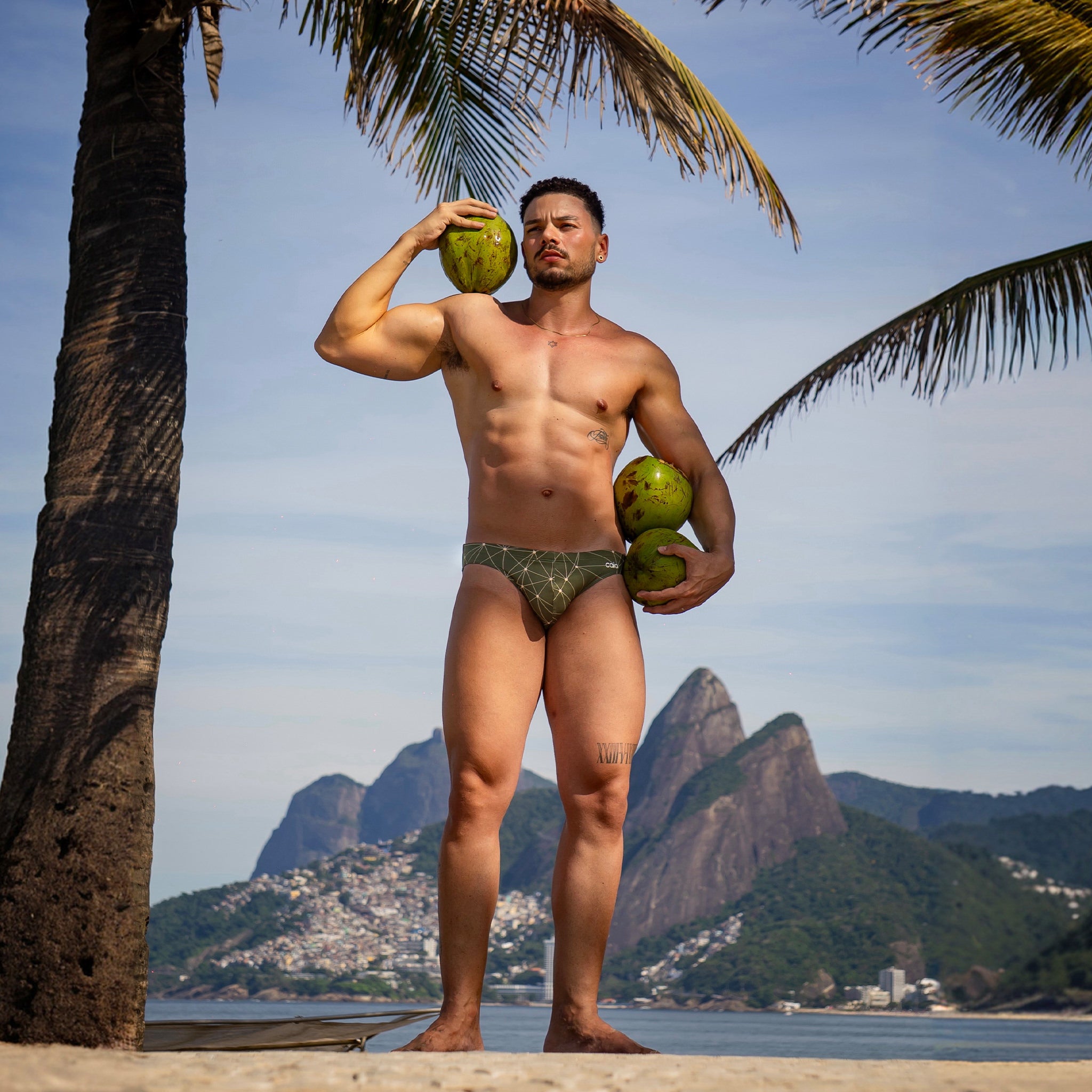 model holding coconuts on a beach with palm trees and mountains in the background wearing caio bikini astro moss