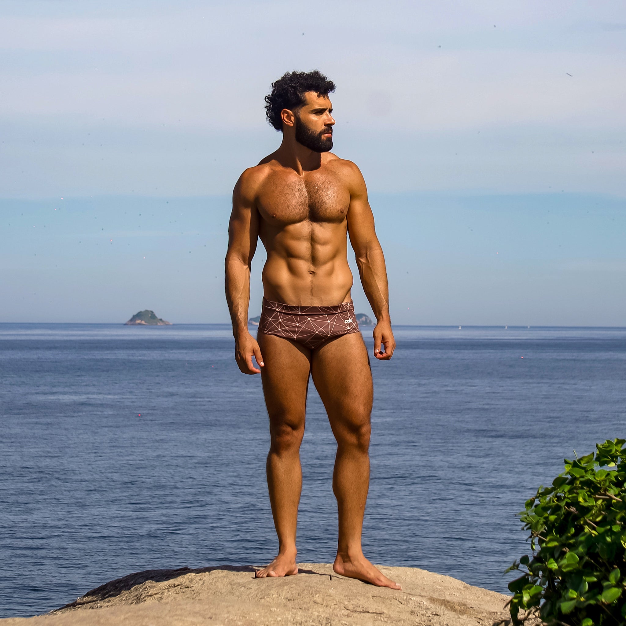 Man standing on a rock by the ocean wearing caio classic astral soil swim brief