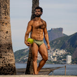 model leaning against palm tree holding coco nut wearing caio slim astro moss swim brief