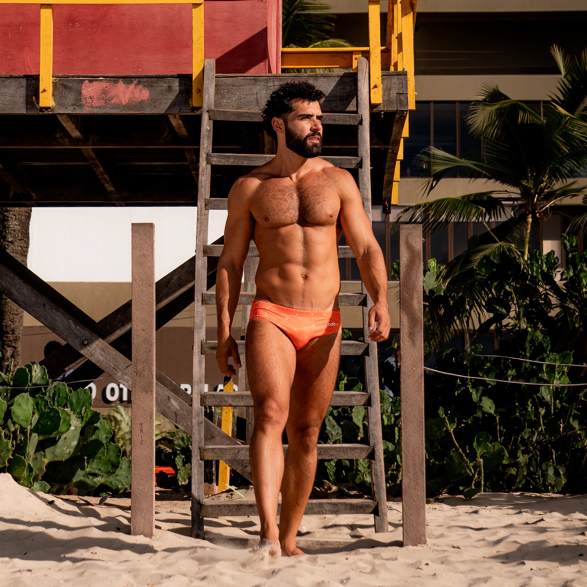Man in Caio Slim Solar Flare swim brief on a sandy beach with a lifeguard tower in the background.