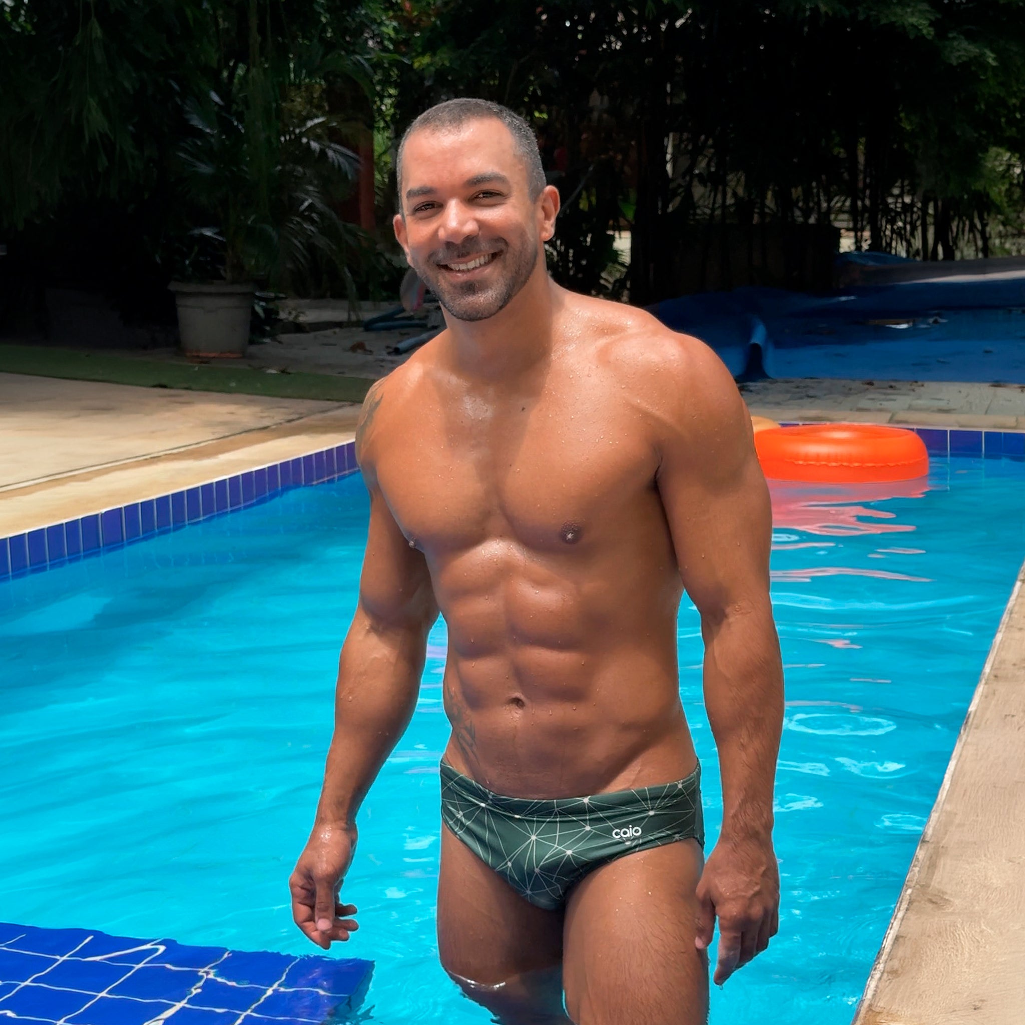 Man standing in a pool wearing caio slim starwood swim brief with a blue tiled pool and orange float in the background.